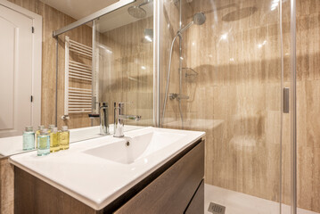 Bathroom with square frameless mirror, white porcelain sink on wooden cabinet, white towel rack, walk-in shower with screen and cream marble cladding on the walls