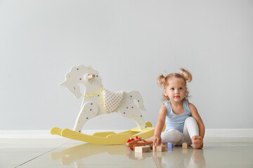 Adorable baby girl with rocking horse and toys near light wall