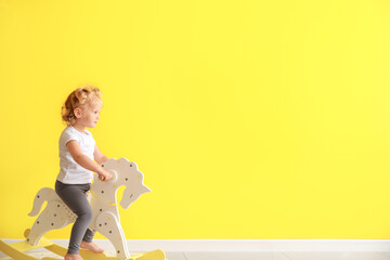 Adorable baby girl with rocking horse near yellow wall