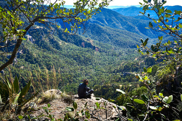 mujer joven sentada frente a un valle con grandes montañas y un hermoso bosque en un día soleado...