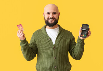 Bald man with payment terminal and credit card on yellow background