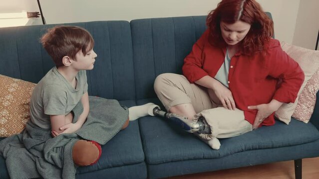 Cheerful happy beautiful disabled 35-40 tutor, teacher woman with prosthetic leg talks to teenage boy child on couch.