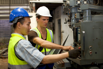 Technician engineer in protective uniform with hardhat standing and teaching apprentices or colleague worker to use computerized machine control at heavy industry manufacturing factory