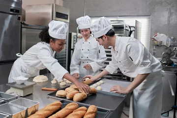 Tuinposter Professional gourmet team, three young chefs in white cook uniforms and aprons knead pastry dough and eggs, prepare bread, and fresh bakery food, baking in oven at stainless steel restaurant kitchen. © tigercat_lpg