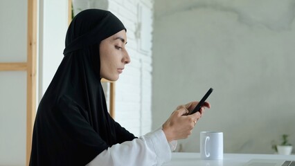 A Muslim woman in a bright cozy room sits and carefully scrolls through the information on the phone. The woman is wearing a hijab. She looks chaste and modest. Engaged in self-development.