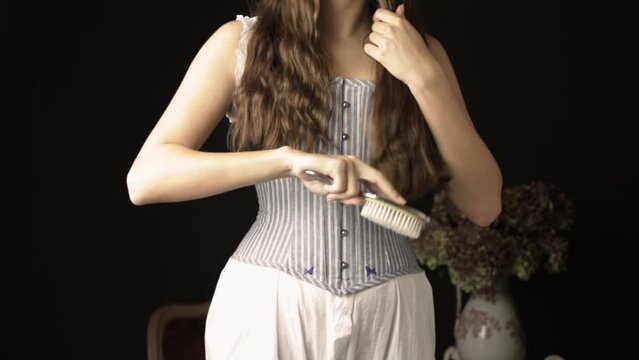 A Victorian woman wearing a corset and combinations brushing her long, dark hair with a silver-backed hairbrush