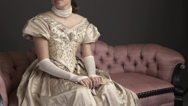 A Victorian woman wearing a gold ball gown with long, fingerless, lace gloves and sitting on a pink sofa in a room resembling a ballroom. A candle light filter has been added.