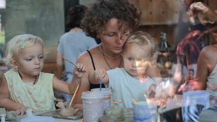Mom with two charming daughters at a master class in clay modeling. Children enthusiastically paint their clay figurines with brushes. Teaching children from an early age craftsmanship.