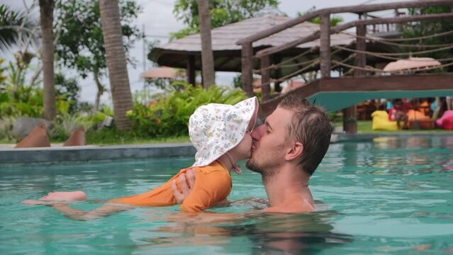 Dad and baby daughter are having fun in the pool, a baby girl in a panama hat and a yellow swimsuit is learning to swim, dad is kissing her daughter. Vacation with children in warm countries.