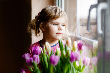 Naklejka premium Little girl sitting by window with tulip flowers bouquet. Happy child, indoors. Mother's day, valentine's day or birthday concept.