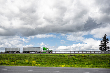 Classic green powerful American big rig semi truck transporting cargo in two bulk semi trailers running on the road on top of the hill