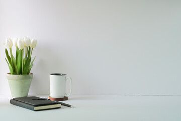 Minimal workplace with white tulips  bouquet in a pot, coffee cup and notebook. Copy space for advertise text or information content.