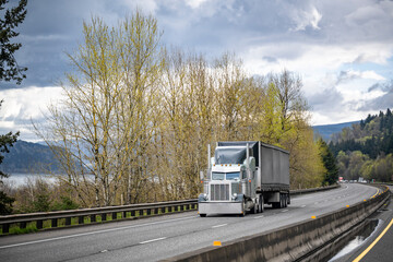 Dark classic American idol big rig semi truck with lot of chrome parts transporting cargo in covered semi trailer moving on the turning highway road along the river