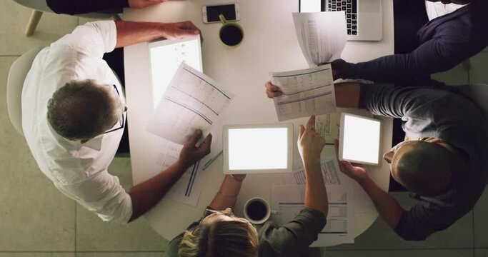 Group of young and diverse business people using tablet technology and reading paperwork while having a meeting in the office