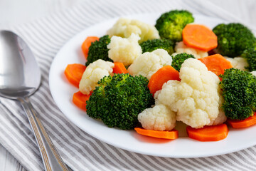 Mixed Organic Steamed Vegetables (Carrots, Broccoli and Cauliflower) on a Plate on a white wooden background, side view. Close-up.
