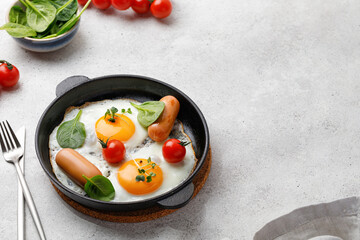 Healthy breakfast is served in a black frying pan. Fried eggs with cherry tomatoes, spinach, sausages.