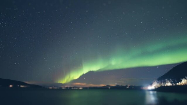 Northern lights above the fjord. Street and car lights flash in the background. Clouds in the sky. Timelapse.
