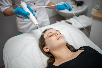Fototapeta premium Beautician uses the darsonval device on the client's face. Professional skin care in the salon. The concept of cosmetology and rejuvenation.