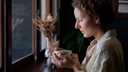 A woman in light clothes, standing at the window, looking out the window, breathing air holds a cup, brings it to her face, inhales the aroma of the freshest drink and is content with this moment.