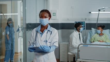 Fototapeta premium Portrait of woman working as doctor with white coat and stethoscope, standing in hospital ward. Medical specialist wearing face mask being ready to treat ill patient with medicine