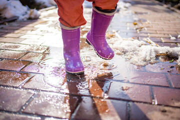 Child jumps on puddles in rubber boots at sunset lights in spring time