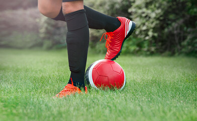 Football training. Child soccer player. Close-up of legs in red turf boots with a beautiful red soccer ball. Foot on the ball.