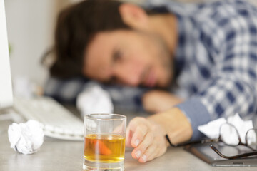 man slumped across desk reaching for alcoholic drink