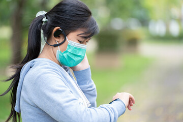 A woman wearing a mask and wearing wireless headphones with a smart watch gets ready for a workout in the park.