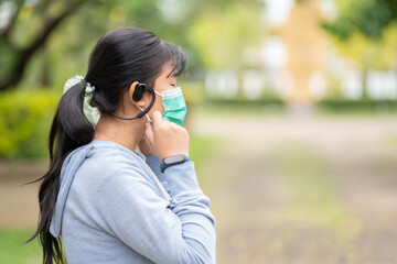 A woman wearing a mask and wearing wireless headphones with a smart watch gets ready for a workout in the park.