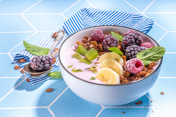 Summer breakfast healthy diet bowl with yogurt, granola, fresh fruit and berries, One portion with overnight fruity granola oats on sun-lighted tiled blue background copy space