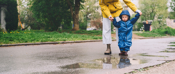 Young mother with her son walking in park in rain. Woman and little child explore nature around playground of residential area in rainy day.
