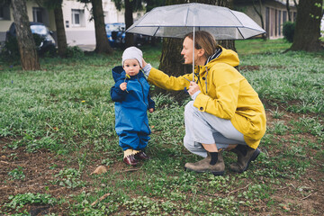 Young mother with her son walking in park in rain. Woman and little child explore nature around playground of residential area in rainy day.