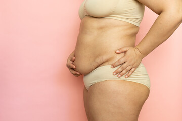 Hold woman sagging belly and stand sideways closeup, folds on stomach, cellulite, obesity. Naked overweight plus size girl on pink background in beige underwear. Concept of dieting and body control.