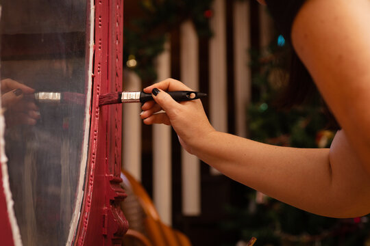 Closeup of woman painting carved ornaments on door with glass of antique cupboard made of wood in pink color. Giving new life to old stuff. Hand restoration of old furniture.