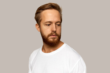 Picture of thoughtful pensive white-skinned bearded male in casual white t-shirt standing against studio wall looking down, trying to remember something important, having stylish haircut