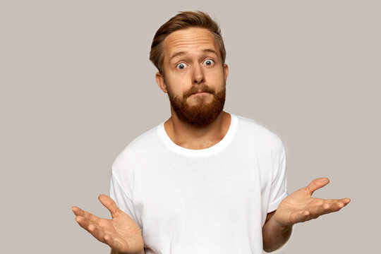 Horizontal picture of confused young bearded man standing and shrugging over grey wall with copy space for your advertisement, making eyes big, throwing hands in the air, looking puzzled