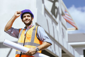 Portrait of a professional heavy industry engineer/worker wearing a safety vest and hard hat smiling on camera in the background. Unfocused large industrial site home.