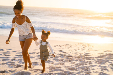 Happy black woman holding hands of daughter while walking on beach at sunset