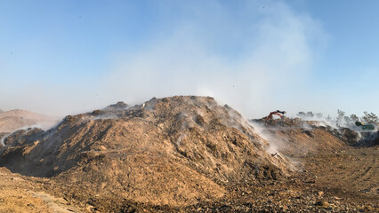 Burning pile of garbage at dump ground or landfill releasing toxic smoke in environment and polluting air. Excavator or earth mover moving or segregating garbage.	