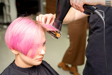 Drying short pink hair of young caucasian woman with a black hairdryer and black round brush by hands of a male hairdresser in a hair salon, close up