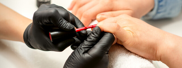 Manicure varnish painting. Close-up of a manicure master wearing rubber black gloves applying red...
