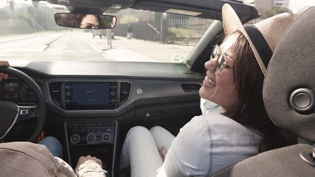 Happy young couple laughing on cabriolet road trip - Smiling woman in summer hat looking her boyfriend while driving a rental car in the city - Vacation and travel concept