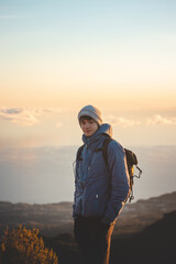 Young adventurer standing on the edge of a rock on the highest mountain Pico Ruivo on the island of Madeira, Portugal at sunrise. The orange-gold light illuminates the young man. Candid portrait