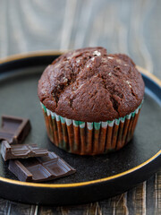 One homemade freshly baked delicious chocolate muffin served on black plate with dark chocolate pieces on table, closeup, side view. Fresh handmade pastry. DIY. Copy space for text. Vertical