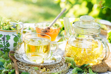 Linden flowers tea in a glass cup and teapot on a white napkin outdoors, yellow drink of linden flower with linden blossoms honey in nature background, healthcare and healthy eating concept