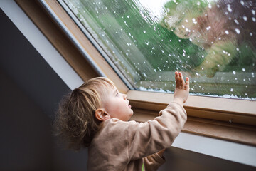 Little preschool boy staying home in bad weather and looking  with interest through window on...
