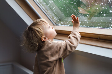 Little preschool boy staying home in bad weather and looking  with interest through window on raindrops and hail, idoors.