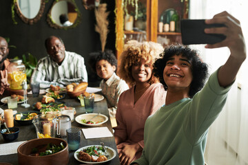 Boy holding mobile phone and making selfie portrait with his family at table during dinner in dining room
