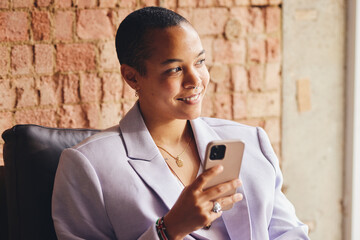 Portrait of multiracial LGBTQ mid adult woman using smartphone and smiling in front of exposed brick wall - obrazy, fototapety, plakaty