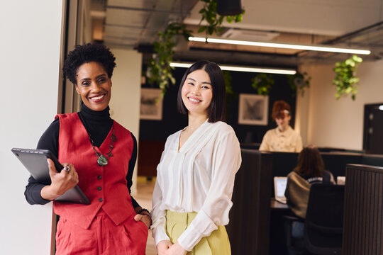 Portrait of cheerful mature black woman and mid adult Chinese female colleague in open plan office space smiling